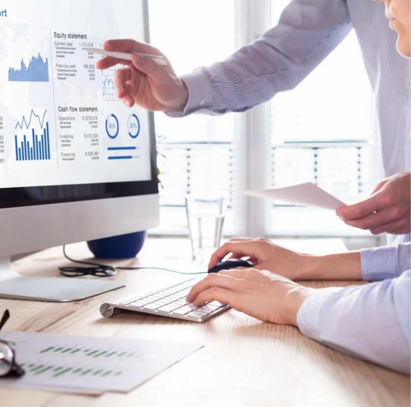 Two people review financial charts and graphs on a computer screen at a desk, with one person pointing and the other using the keyboard.